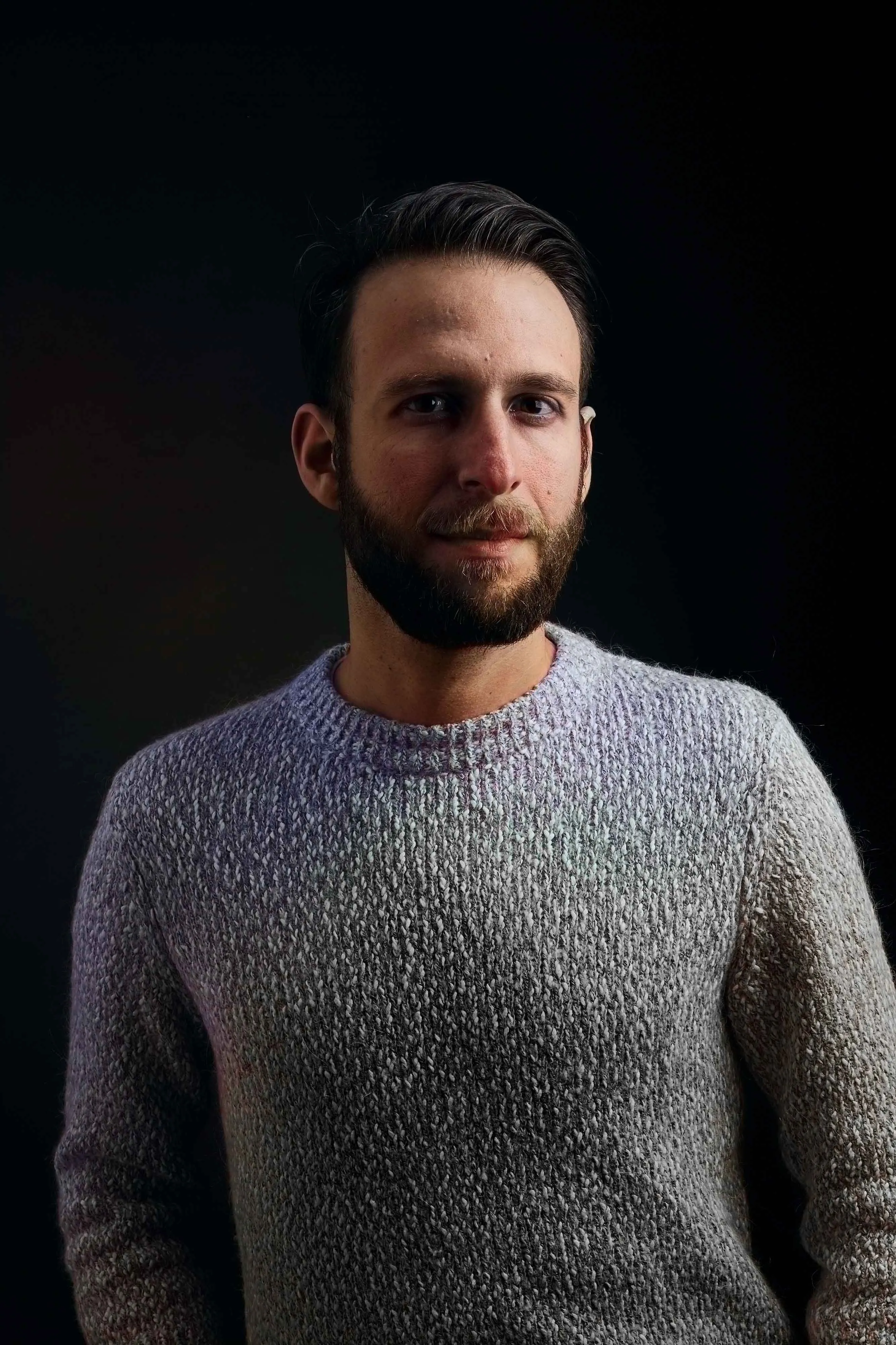 Portrait of a man with dark hair and beard wearing a textured gray sweater against a dark background.
