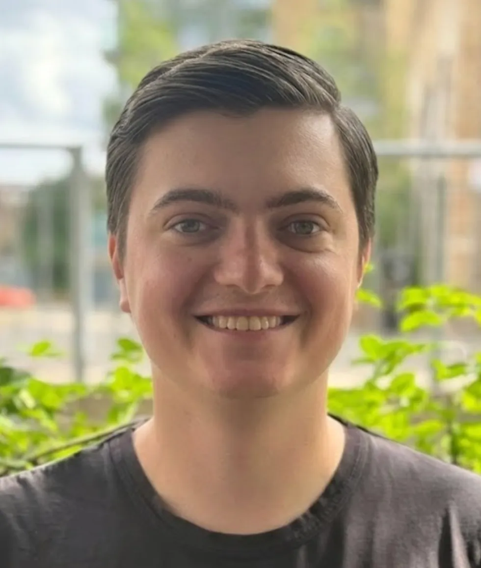 Smiling young man with short dark hair and light skin wearing a dark t-shirt, with green plants and an outdoor blurred background.