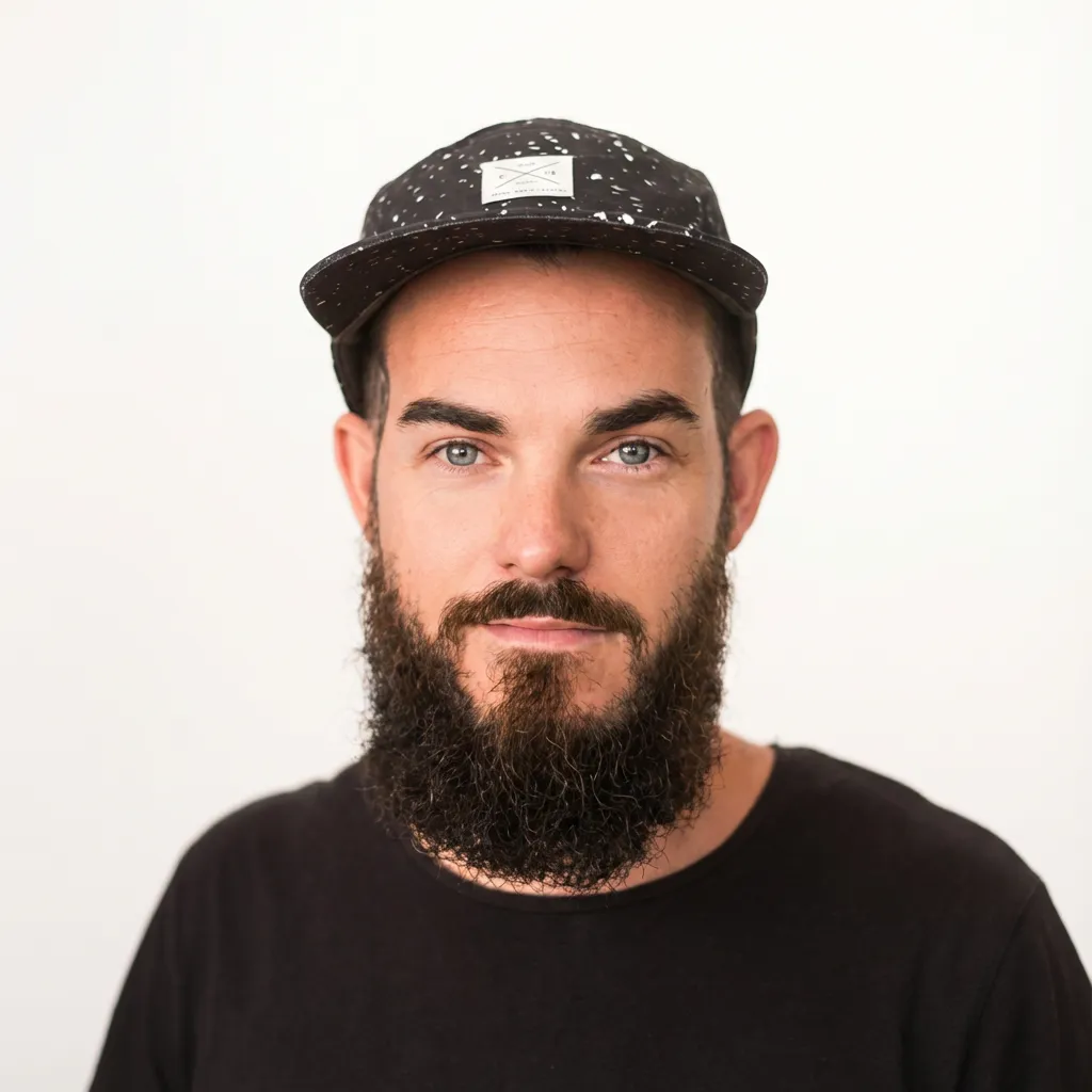 Portrait of a man with a thick dark beard and mustache, light eyes, wearing a black speckled cap and black shirt, against a plain white background.