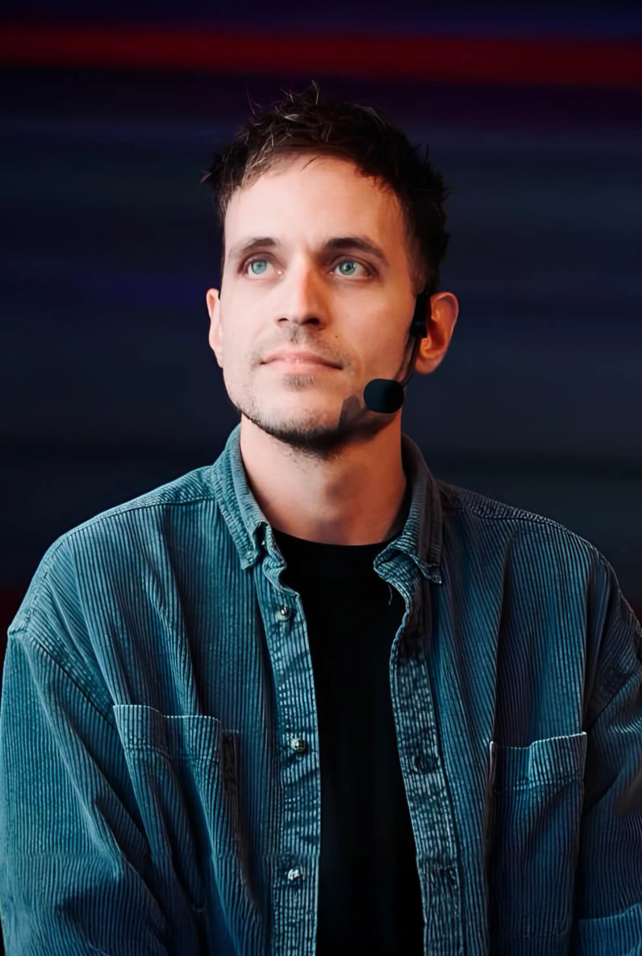 Young man with light skin and blue eyes wearing a dark blue corduroy shirt over a black T-shirt, looking slightly upward and to the left, with a headset microphone on his right ear against a dark blurred background.