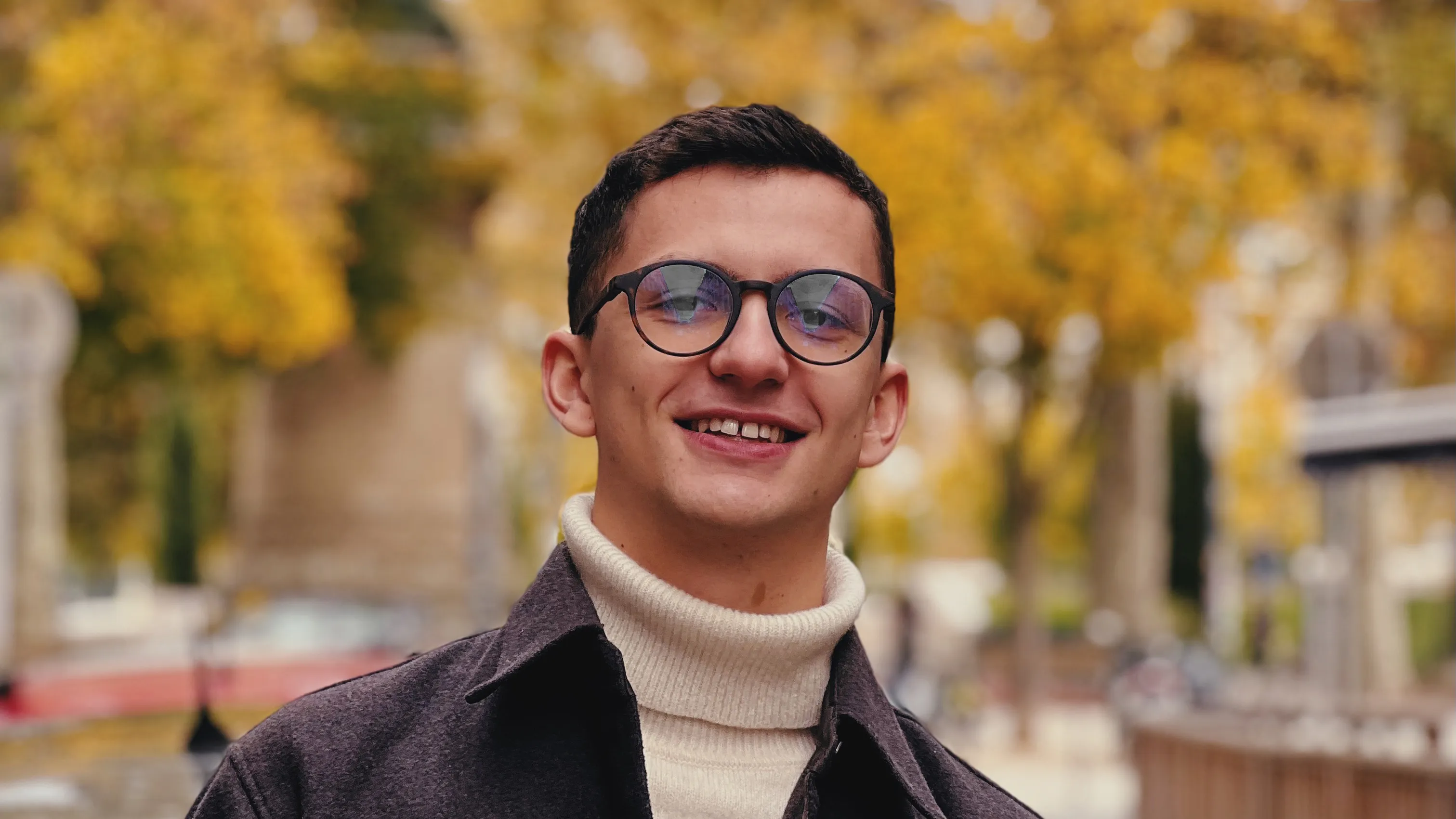 Young man with short dark hair and round glasses smiling while wearing a white turtleneck sweater and dark coat, with blurred autumn trees in the background.