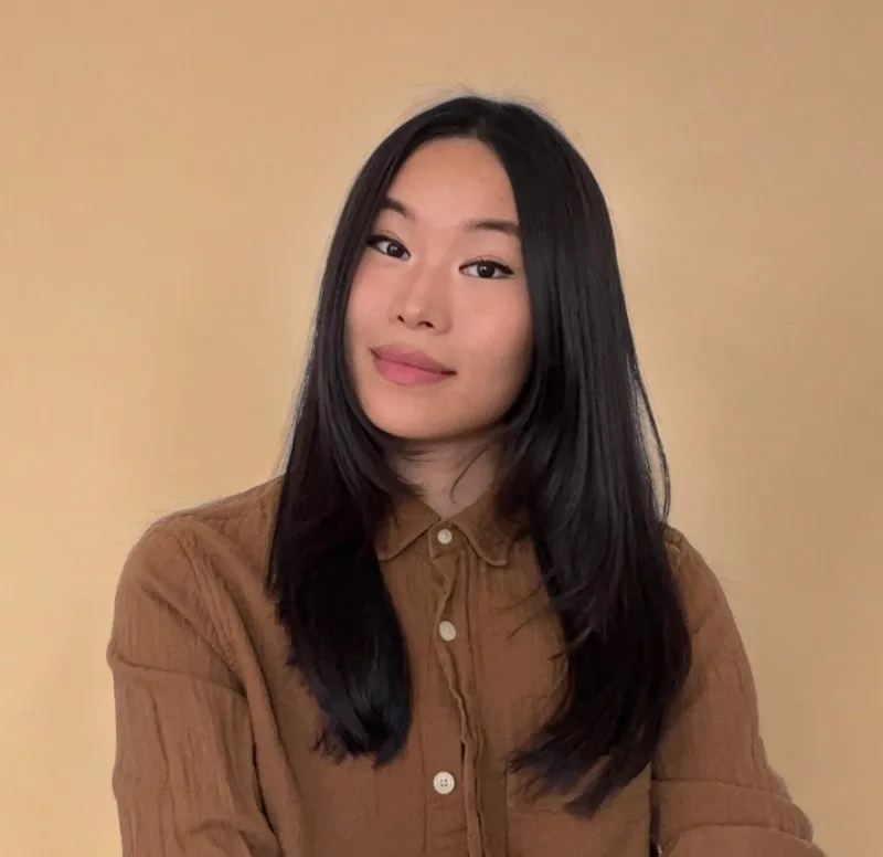 Portrait of a young woman with long black hair wearing a brown button-up shirt, posing against a plain beige background.