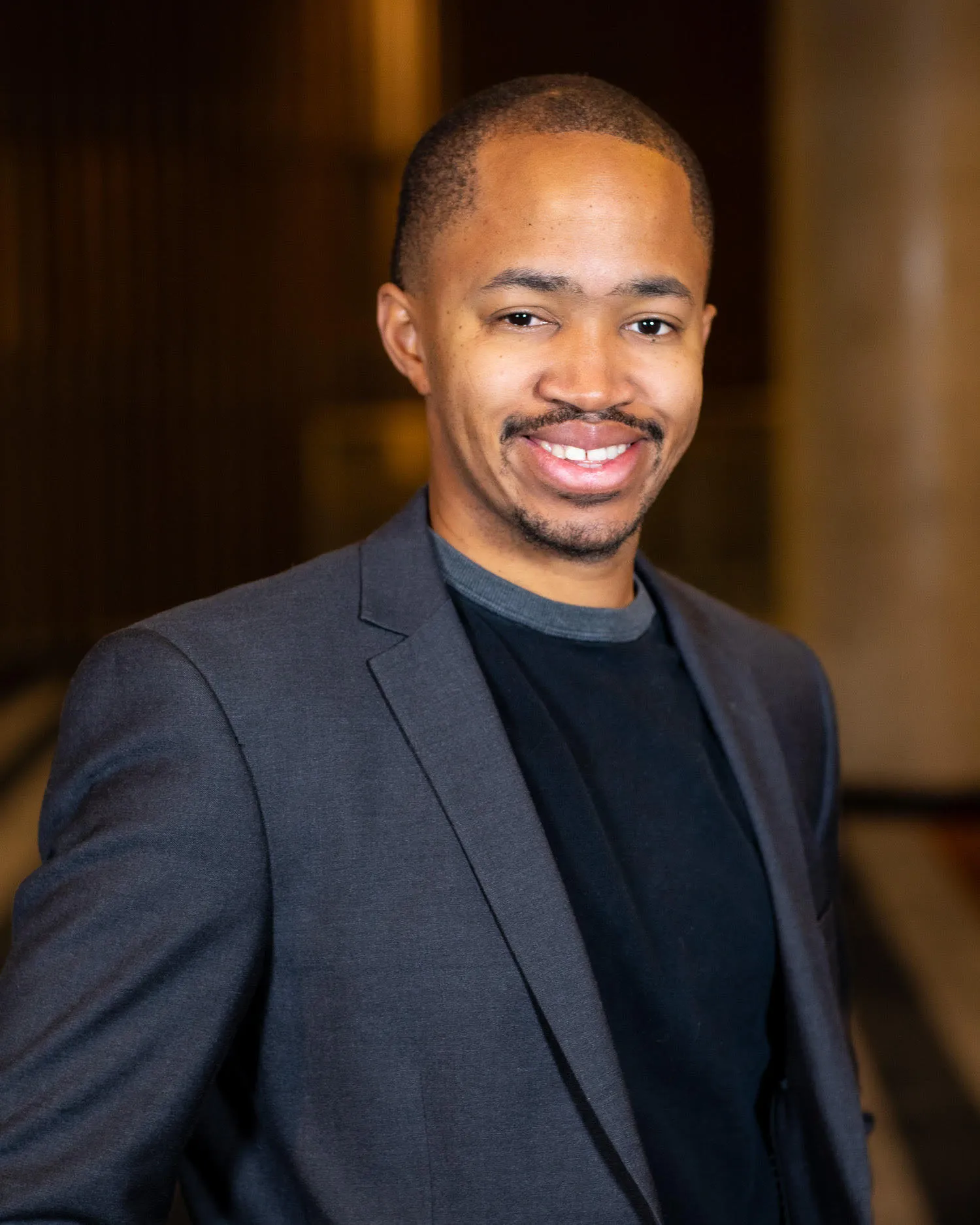 Smiling man wearing a dark gray blazer and black shirt stands against a blurred indoor background.